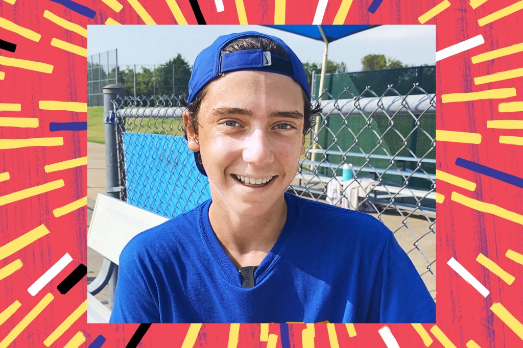 boy in blue cap and shirt smiling at a fence at a sports field